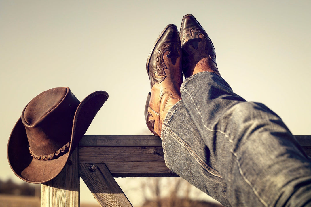 Southern living - person's legs propped up on a fence wearing cowboy boots with a cowboy hat sitting on the edgs of the fence post