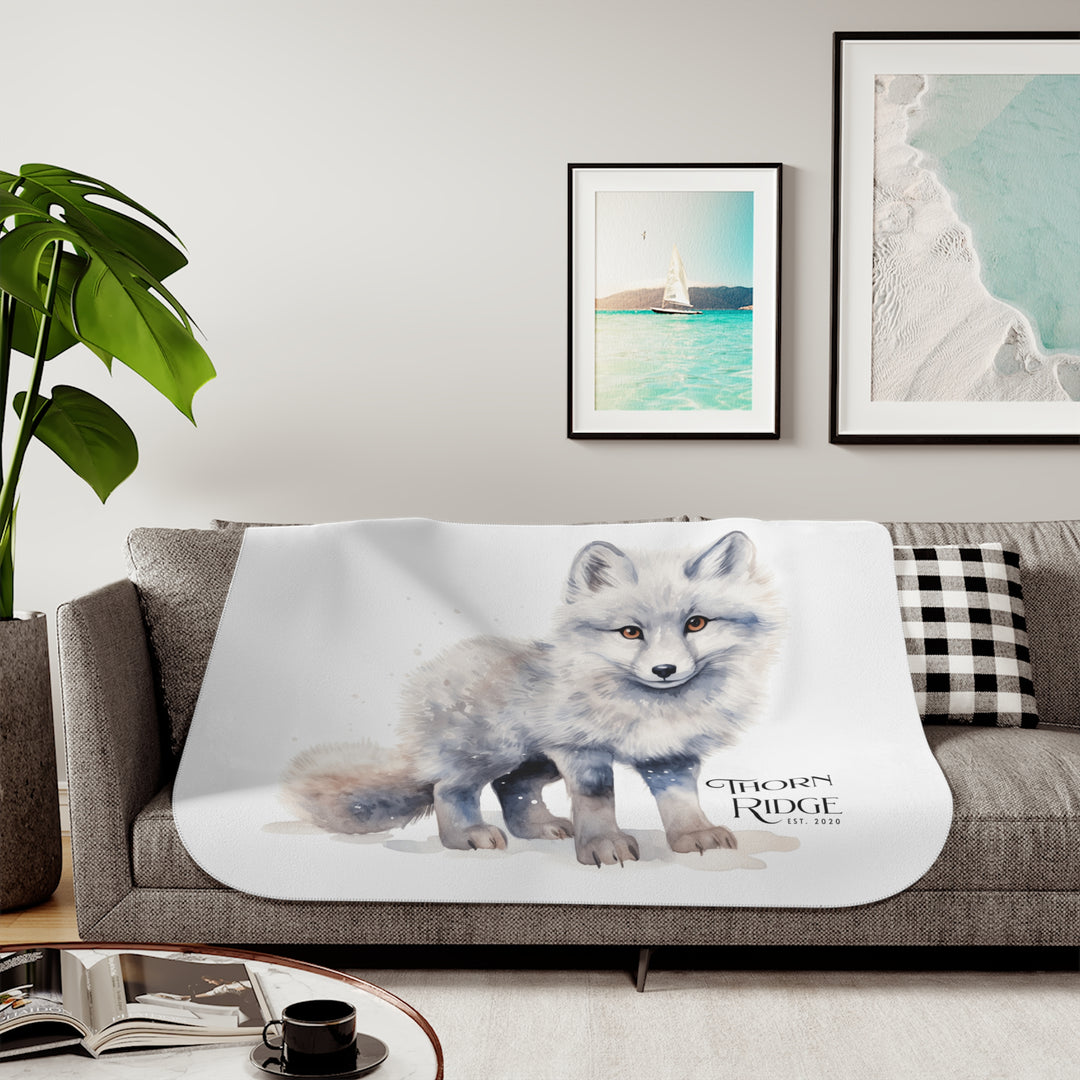 White Arctic fox illustration on a white Sherpa Blanket with 'Thorn Ridge' branding and the blanket is laying across a couch. 