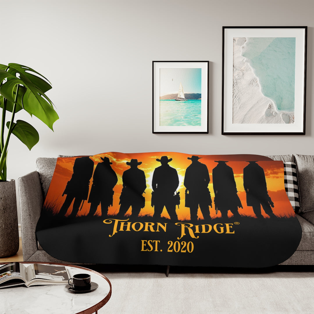 Silhouette of Seven Riders cowboys against an orange sunset with 'Thorn Ridge' branding on a sherpa blanket on a couch.