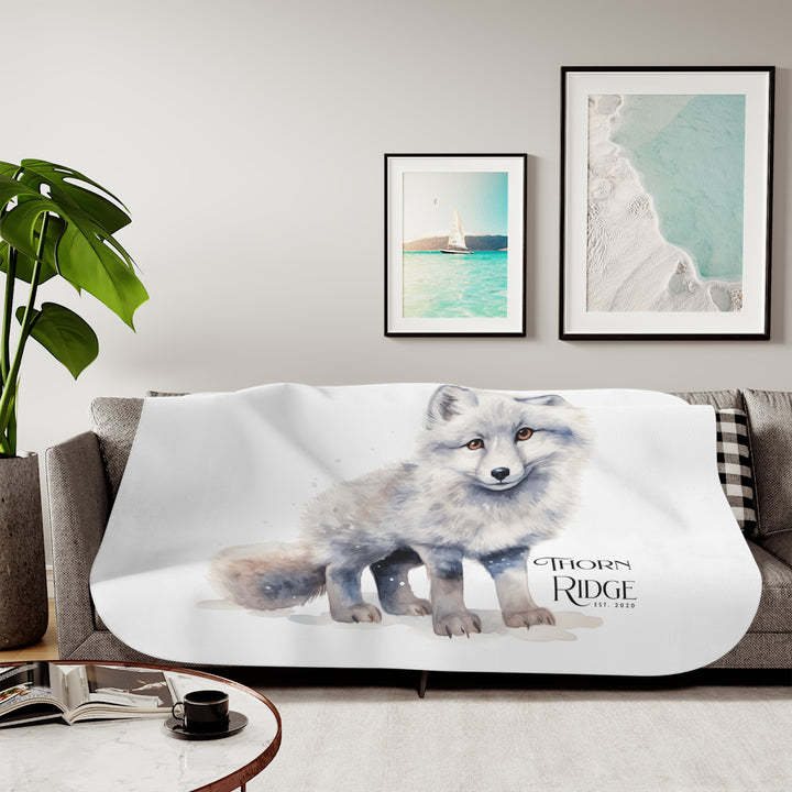 White Arctic fox illustration on a white Sherpa Blanket with 'Thorn Ridge' branding and the blanket is laying across a couch. 