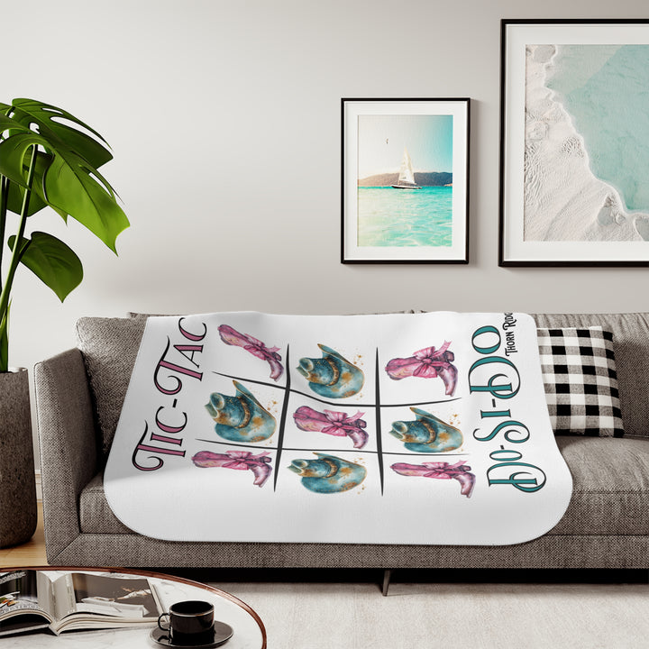 White Sherpa Blanket with a Tic-Tac Do-Si-Do image of a tic-tac-toe board with pink cowboy boots with a bow in the four corners and middle and blue cowboy hats on the sides by Thorn Ridge®