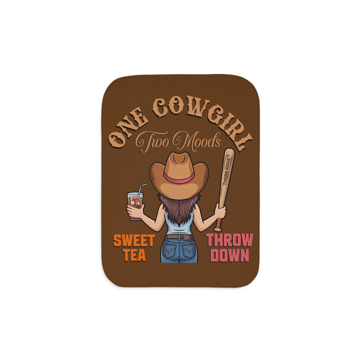 One Cowgirl Two Moods Sherpa Blanket by Thorn Ridge that's brown with a illustration of a cowgirl from the back holding a glass of sweet tea in the left hand and a baseball bat labeled Thorn Ridge® in the right hand with the words sweet tea under the glass and throw down under the bat. 