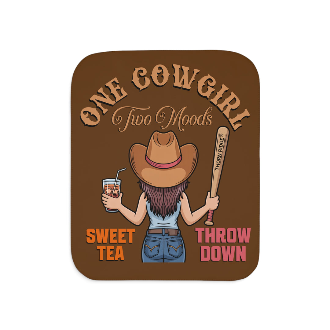 One Cowgirl Two Moods Sherpa Blanket by Thorn Ridge that's brown with a illustration of a cowgirl from the back holding a glass of sweet tea in the left hand and a baseball bat labeled Thorn Ridge® in the right hand with the words sweet tea under the glass and throw down under the bat. 