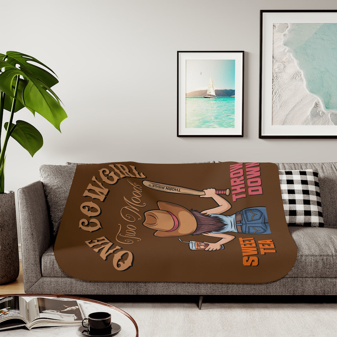 One Cowgirl Two Moods Sherpa Blanket by Thorn Ridge that's brown with a illustration of a cowgirl from the back holding a glass of sweet tea in the left hand and a baseball bat labeled Thorn Ridge® in the right hand with the words sweet tea under the glass and throw down under the bat. 