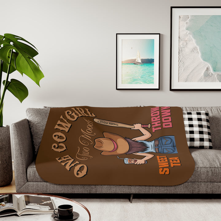 One Cowgirl Two Moods Sherpa Blanket by Thorn Ridge that's brown with a illustration of a cowgirl from the back holding a glass of sweet tea in the left hand and a baseball bat labeled Thorn Ridge® in the right hand with the words sweet tea under the glass and throw down under the bat. 