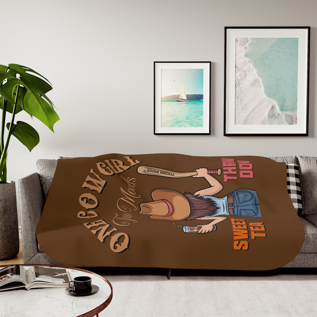 One Cowgirl Two Moods Sherpa Blanket by Thorn Ridge that's brown with a illustration of a cowgirl from the back holding a glass of sweet tea in the left hand and a baseball bat labeled Thorn Ridge® in the right hand with the words sweet tea under the glass and throw down under the bat. 