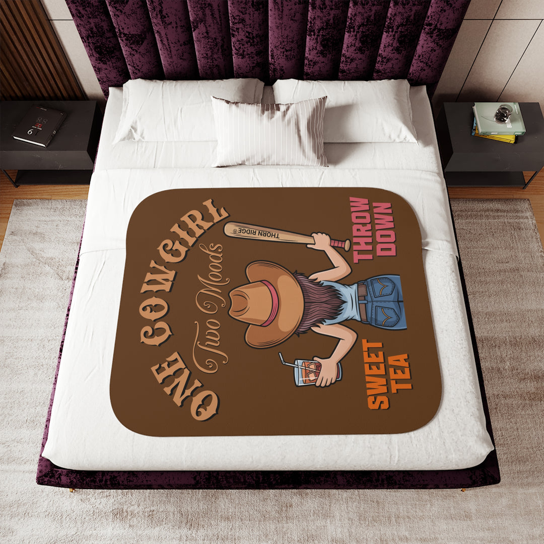 One Cowgirl Two Moods Sherpa Blanket by Thorn Ridge that's brown with a illustration of a cowgirl from the back holding a glass of sweet tea in the left hand and a baseball bat labeled Thorn Ridge® in the right hand with the words sweet tea under the glass and throw down under the bat. 