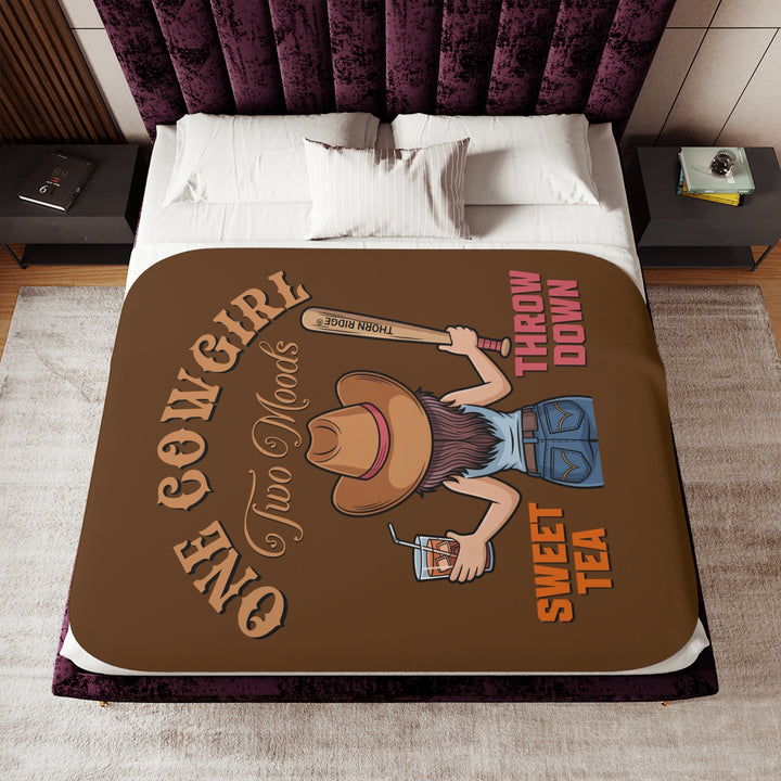 One Cowgirl Two Moods Sherpa Blanket by Thorn Ridge that's brown with a illustration of a cowgirl from the back holding a glass of sweet tea in the left hand and a baseball bat labeled Thorn Ridge® in the right hand with the words sweet tea under the glass and throw down under the bat. 