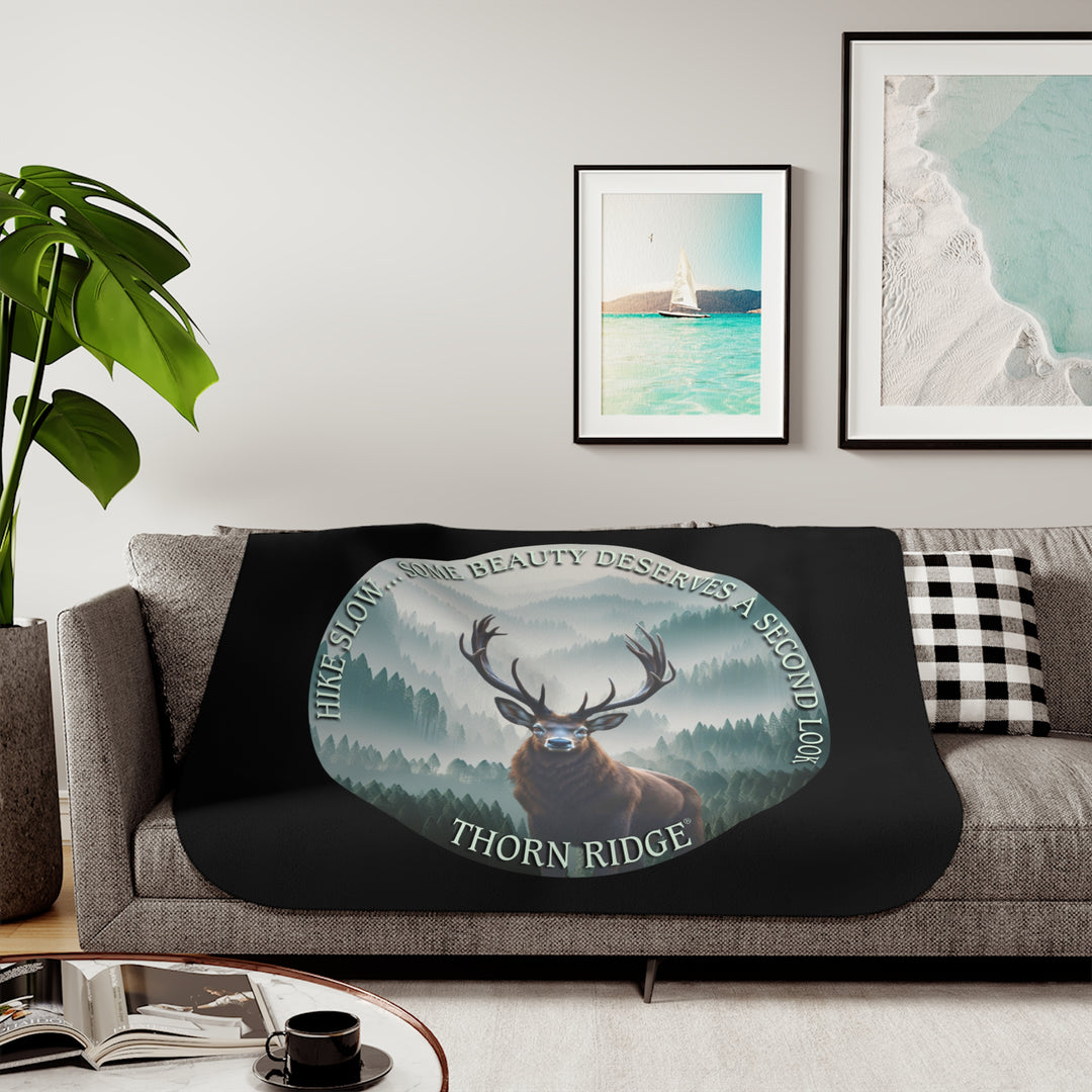 Hike Slow some beauty deserves a second look black sherpa blanket featuring a circular graphic of a buck looking at the camera with a green misty forest behind him and branded Thorn Ridge®