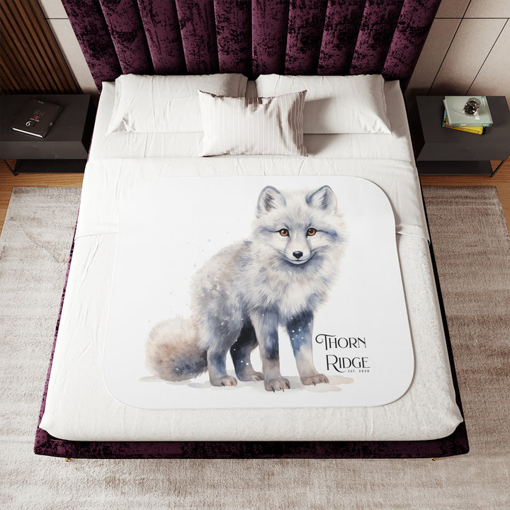 White Arctic fox illustration on a white Sherpa Blanket with 'Thorn Ridge' branding and the blanket is laying across a bed. 