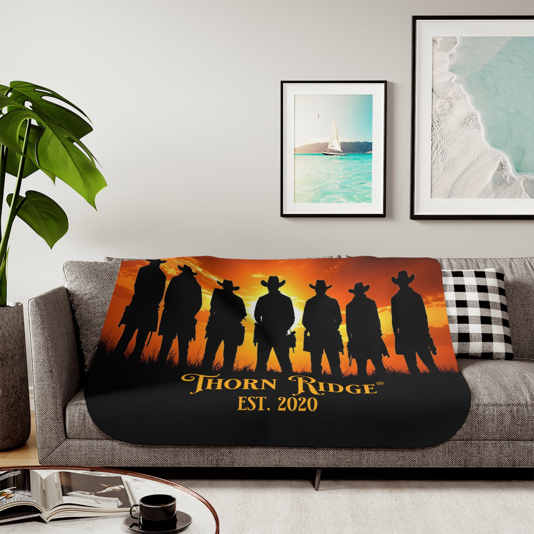 Silhouette of Seven Riders cowboys against an orange sunset with 'Thorn Ridge' branding on a sherpa blanket on a couch.